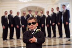 a young boy wearing sunglasses and standing in front of a group of men with their arms crossed