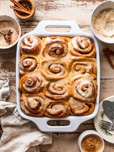 a baking dish filled with cinnamon rolls on top of a wooden table next to bowls and utensils