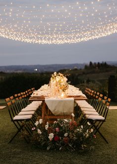 an outdoor dinner table with lights strung from the ceiling and flowers on the table, surrounded by greenery