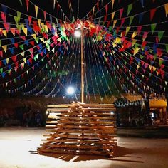 a large pile of wood sitting in the middle of a yard at night with colorful flags hanging above it