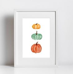 three watercolor pumpkins on white paper in front of a white wall and shelf