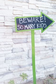 a black and green sign that says beware so many kids next to a brick wall