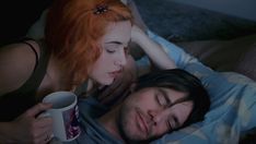 a man and woman laying on top of a bed next to each other holding coffee mugs