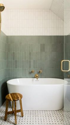 a white bath tub sitting next to a wooden stool in a bathroom with green tiles on the walls