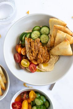 two bowls filled with different types of food on top of a white countertop next to french fries and cucumbers