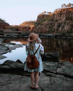 a woman in a dress and hat standing on rocks looking out at the water with her back to the camera