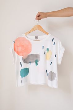 "Cropped organic cotton T-shirt, with round neck, white base and hand painted motifs in bright tones (salmon, mustard, blue green, gray, blue). This shirt is made in Africa with ethical craftsmanship. The cropped shape with rolled sleeves, combined with the manual water stains, make it perfect for both relaxed and sophisticated looks. You'll look radiant with a high-waisted jean or with dress pants and a jacket, achieving the \"effortless elegance\" effect that we love at APPA Studio. The \"Colo Salmon Mustard, Color Cloud, Cloud Print, Water Stains, Effortless Elegance, Clothing Care, How To Make Tshirts, High Waist Jeans, Tshirt Colors