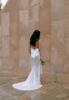 a woman in a white dress is standing near a stone wall and holding a bouquet