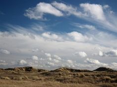 there are clouds in the blue sky above some brown hills and grass on the ground