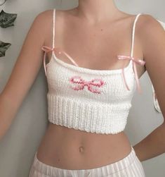 a woman wearing a white crop top with pink bows on the front and side, standing next to a wall