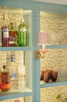 bottles and glasses sit on shelves in a room with floral wallpaper, one is green and the other is blue