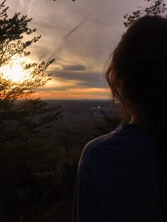 a woman looking out at the sunset with her back to the camera and trees in the foreground