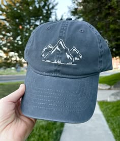 a person holding up a hat with mountains on it in front of grass and trees