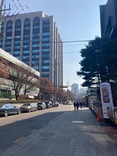 people are walking down the street in front of some tall buildings and cars parked on the side of the road