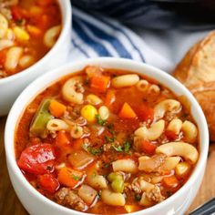 two white bowls filled with pasta soup on top of a cutting board next to bread