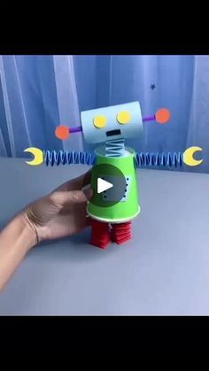 a hand holding a green and red robot toy on top of a gray table next to a blue curtain