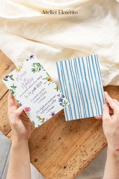 two hands holding an open wedding card on top of a wooden table with white and blue striped paper