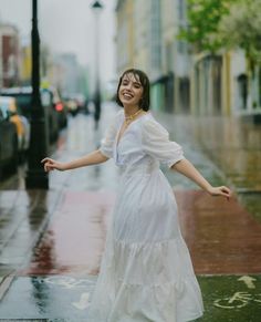 a woman in a white dress is standing on the sidewalk with her arms spread out