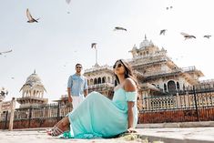 a man and woman sitting on the ground in front of a building with birds flying around