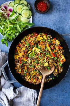 a skillet filled with rice and vegetables next to a bowl of cucumbers