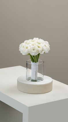 a white flower arrangement in a glass vase on top of a white table with a gray wall behind it