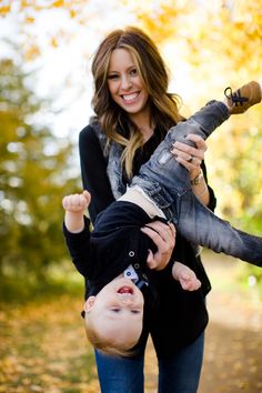 a woman holding a baby in her arms and smiling at the camera with autumn leaves on the ground