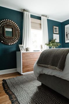 a bedroom with teal walls and white drapes on the windowsills, a gray bed in front of a black and white patterned rug