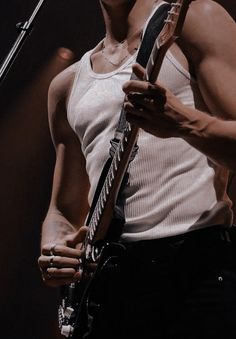 a man holding a guitar while standing in front of a microphone and wearing a white tank top