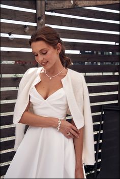 a woman in a white dress and jacket standing next to a wooden fence with her hand on her hip