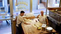 two men standing in front of a wooden table with food on it and one man pointing at the camera