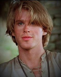 a young man with blonde hair and blue eyes looks at the camera while wearing a necklace