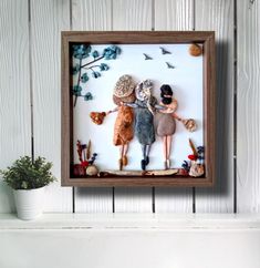 two dolls are holding hands in a shadow box on a shelf next to a potted plant