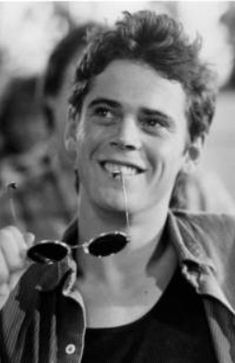 a black and white photo of a smiling man holding two spoons in his mouth