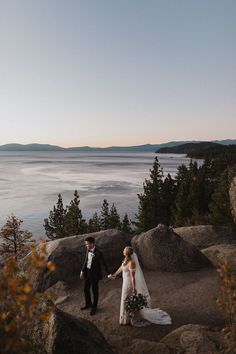 a bride and groom holding hands while standing on top of a cliff overlooking the ocean