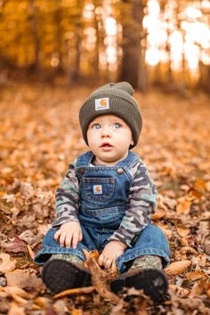 a little boy sitting in the leaves wearing a hat