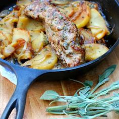 a skillet filled with meat and potatoes on top of a wooden table next to sage