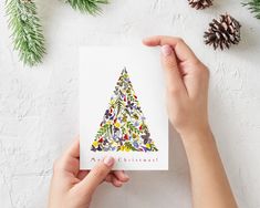 someone holding up a card with a christmas tree on it, surrounded by evergreen branches and pine cones