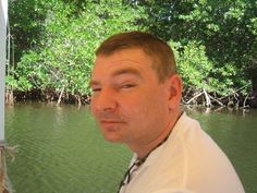 a man standing in front of a body of water with trees behind him and looking at the camera