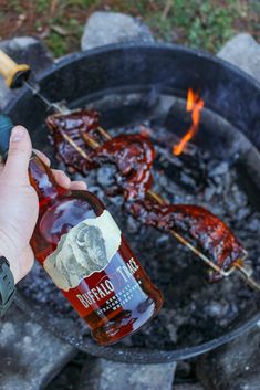 a person is holding a bottle over an open fire pit with hot dogs on it