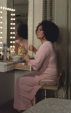 a woman sitting at a vanity in front of a mirror with lights on the wall