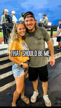 a man and woman standing next to each other in front of a baseball field with the words that should be me