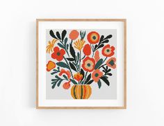 a cross stitch pattern with an orange vase filled with flowers on a white wall next to a wooden frame