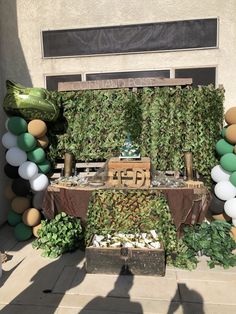 a table topped with lots of green and white balloons next to a wall covered in greenery