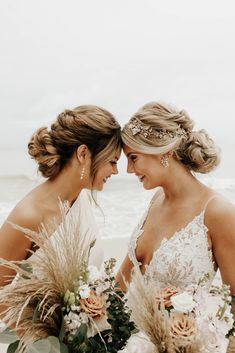 two women standing next to each other holding bouquets in front of the ocean on a cloudy day