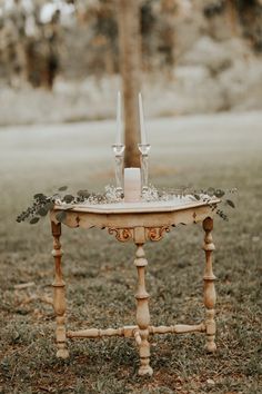 an old table with two candles on it in the middle of a grassy area next to a tree