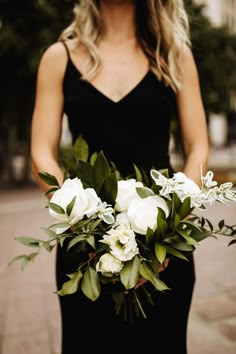 a woman in a black dress holding a bouquet of white roses and greenery on the street