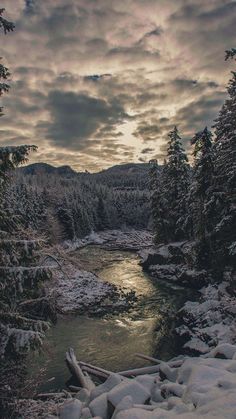 the river is surrounded by snow covered trees and mountains in the distance, under a cloudy sky