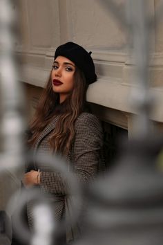 a woman leaning against a wall wearing a black hat and coat with her arms crossed