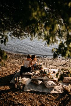 a man and woman sitting on a blanket at the beach with food in front of them