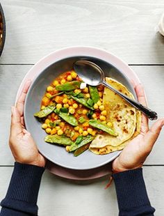 two hands holding a plate of food with corn and tortillas on the side